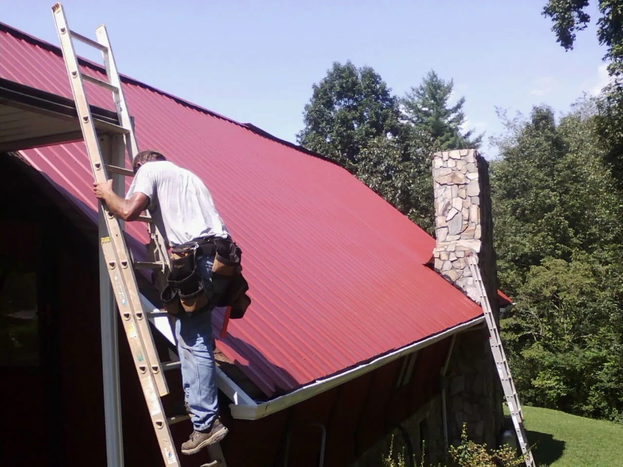 Roofer on a ladder beside a red metal roof and stone chimney in Narrowsburg